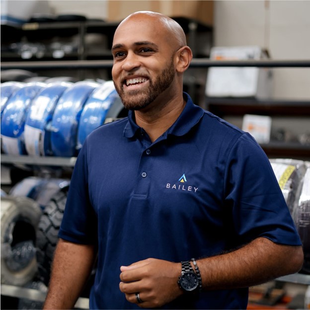 Person wearing a navy blue Bailey-branded polo shirt and a wristwatch standing in an industrial setting with shelves of tires and equipment in the background.