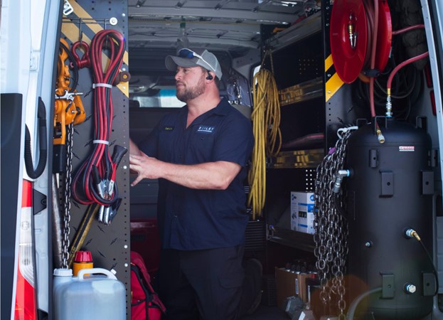 Person wearing a dark Bailey-branded work shirt and cap standing inside a service van organized with tools and equipment. The van contains coiled hoses, chains, a large black air tank, and various maintenance supplies neatly arranged on shelves.